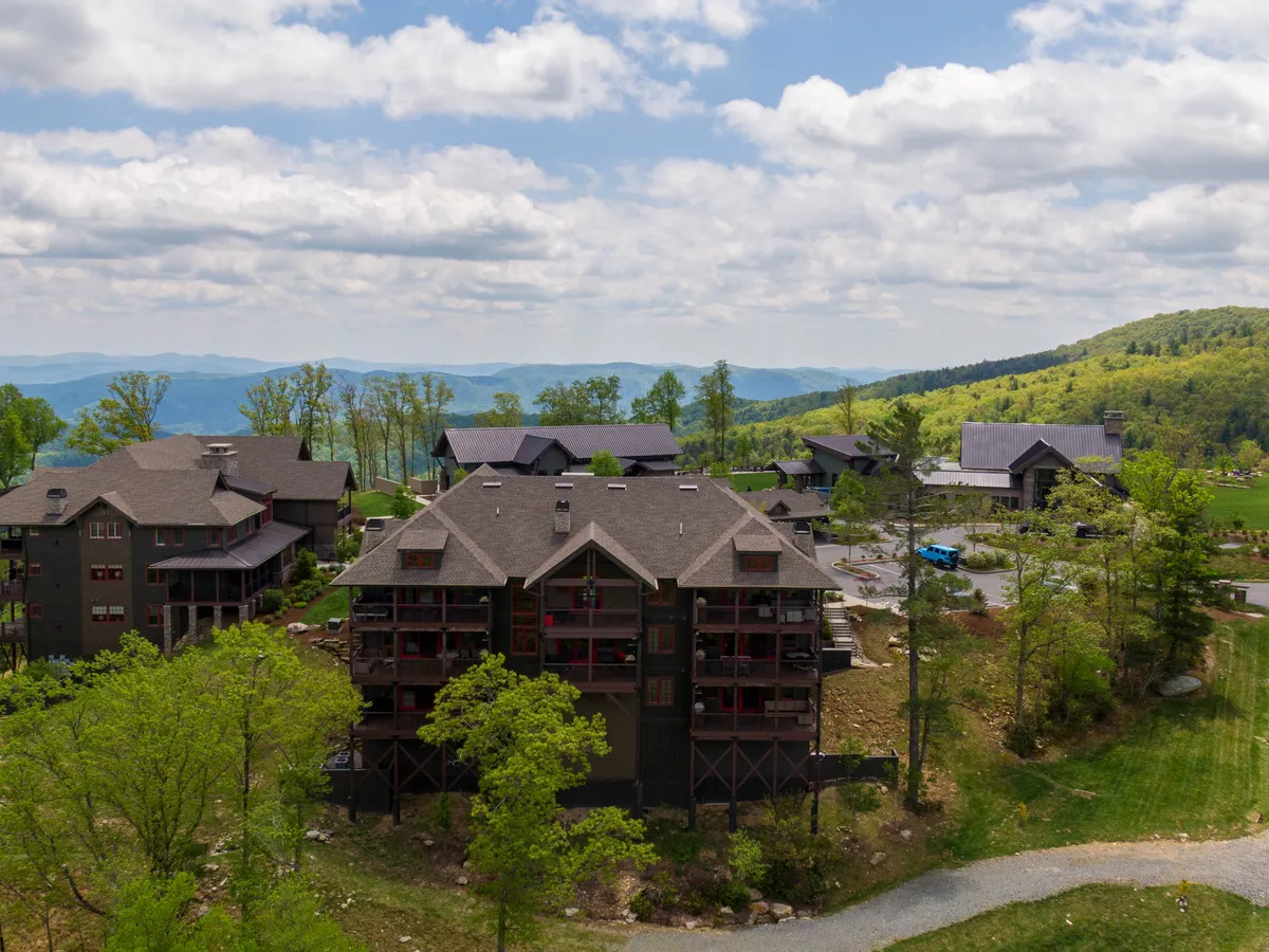 This image captures an inviting exterior scene with scenic mountain views, wooden architecture, lush green trees, and a clear blue sky. The setting evokes a sense of exploration and connection to nature, with the landscape offering a glimpse of the expansive outdoors available to residents and guests.