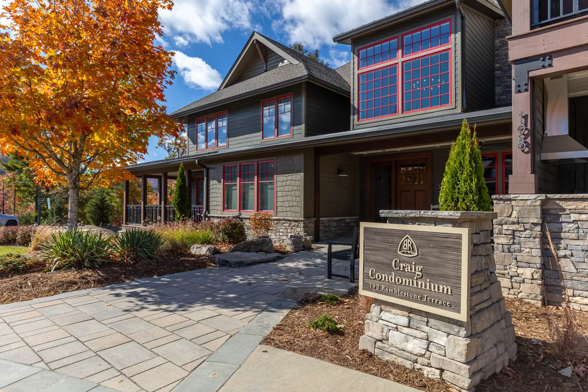 The image showcases the exterior of the Craig Condominium building at the Blue Ridge Mountain Club during vibrant autumn. The welcoming stone facade and distinctive architecture are framed by a brilliant red-orange fall tree, with a visible sign marking 'Craig Condominium.' The inviting porch and natural surroundings hint at the relaxing, nature-immersed experience awaiting guests.