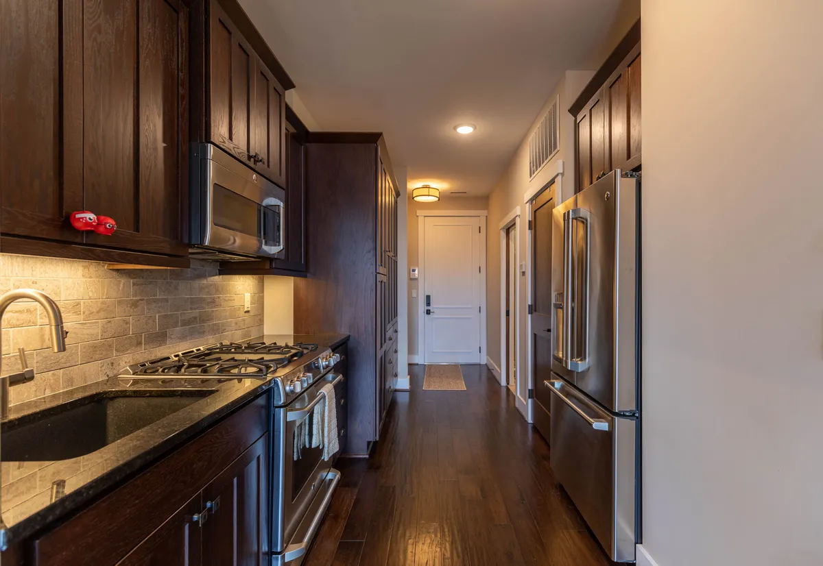 A warmly lit kitchen featuring dark wood cabinetry, modern kitchen appliances, and a granite countertop. The kitchen flows into a hallway, suggesting openness and ease of movement. The space feels inviting and is clearly designed for cooking and entertaining.