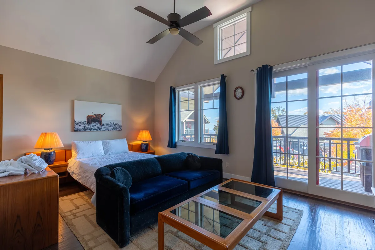 This image captures a tranquil studio space featuring a plush queen bed, a cozy sofa, a large window letting in abundant natural light, and tasteful artwork adorning the walls. The soft lighting and uncluttered layout create a welcoming, restful atmosphere ideal for relaxation.