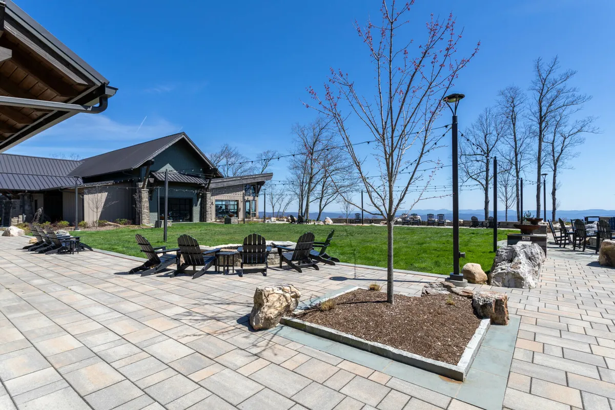 An outdoor seating area shaded by a tree, adjacent to a landscaped lawn and the facade of a building. The setting features a peaceful, communal fire pit area ideal for evening gatherings or quiet moments shared between couples.
