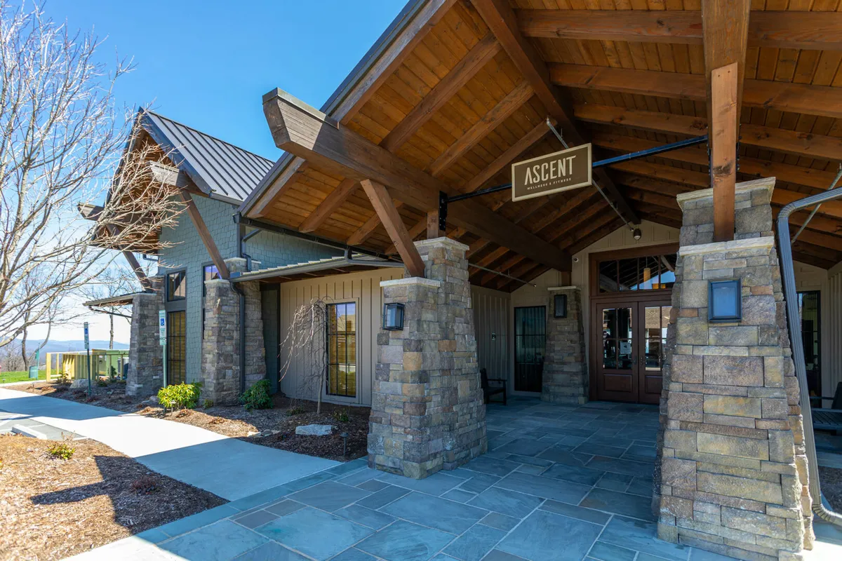The welcoming entrance to the Ascent Wellness & Fitness Center, featuring an inviting sign, sturdy stone pillars, wooden beams, and a landscaped garden. The tranquil atmosphere hints at the wellness amenities within.