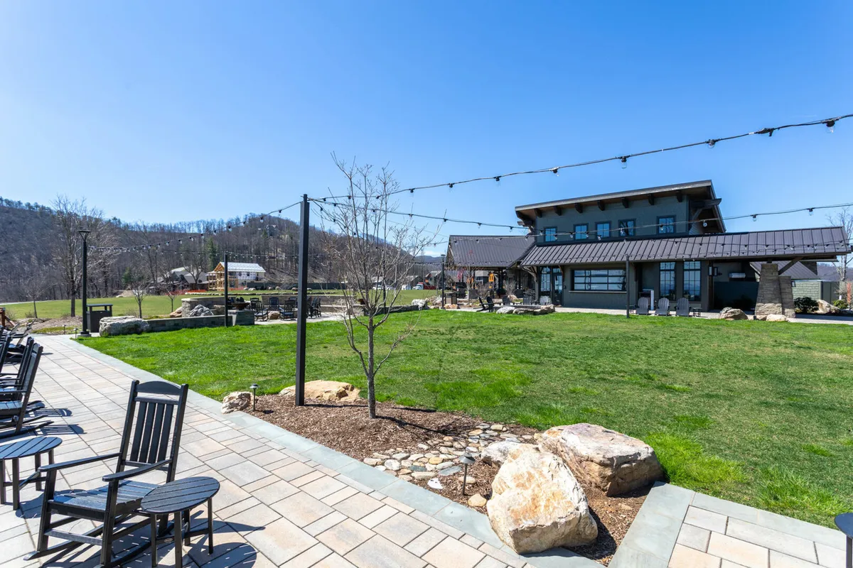 The photo features an expansive lawn with rocking chairs, a modern building in the background, and strings of lights overhead. This communal outdoor space is ideal for gatherings, relaxing with friends, or playing outdoor games, echoing the community-focused amenities described in the section.