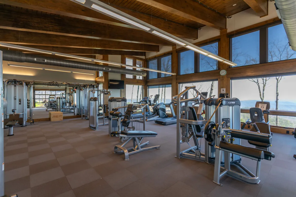 A bright, modern fitness center interior with exercise machines, large windows offering scenic mountain views, and exposed wooden beams. The space is well-lit, inviting, and designed for personal training, group classes, or solo workouts.